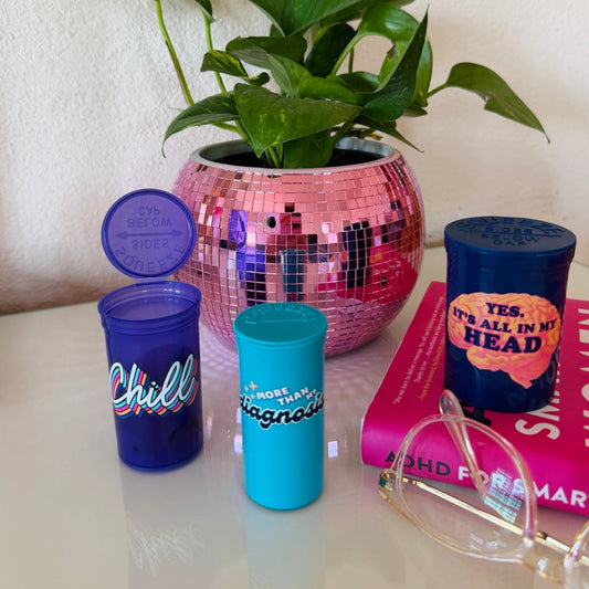 Three colorful Happy Pills pill bottles displayed besides a pink disco ball planter, books and glasses. 